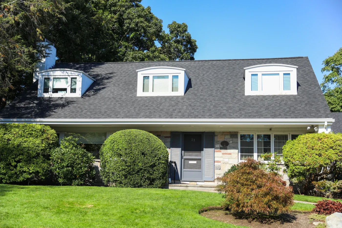 A multi-colored brick house with dormer windows and an asphalt shingle roof with bushes in front.