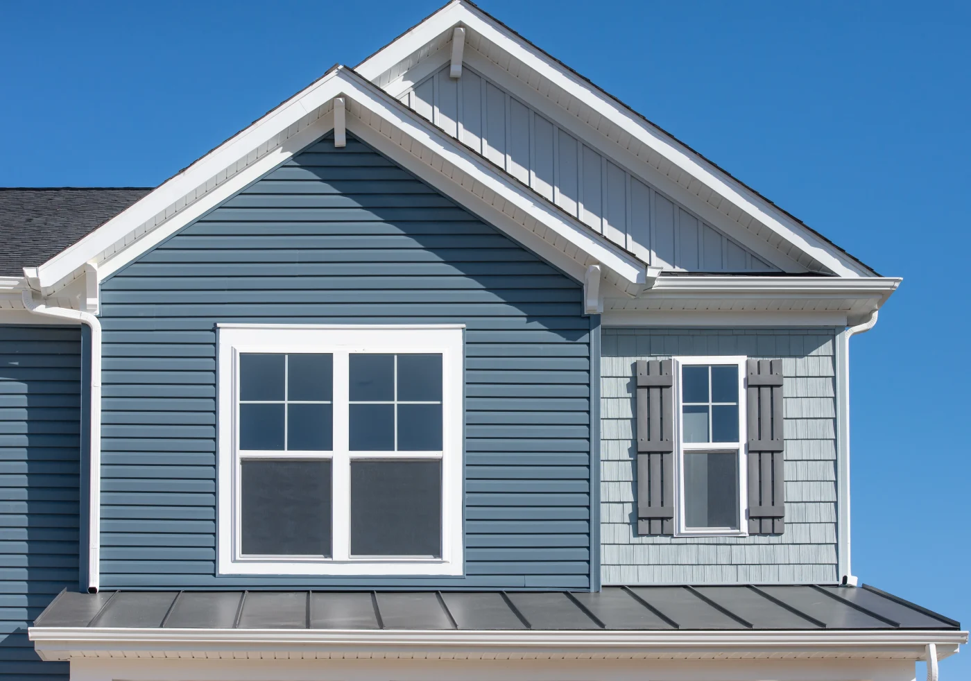 Elegant Blue Horizontal Vinyl Siding, Shingle And Vertical Siding In A Double Gable Roof With White Decorative Corbels, Double Sash Window With Matching Shutter Above A Metal Roof On A Luxury Home Elegant Blue Horizontal Vinyl Siding, Shingle And Vertical Siding In A Double Gable Roof With White Decorative Corbels, Double Sash Window With Matching Shutter Above A Metal Roof On A Luxury Home