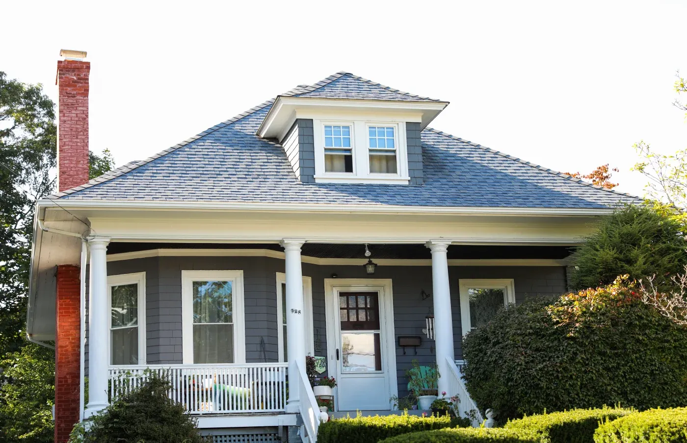 A gray house with a white porch and an asphalt shingle roof with a brick chimney.