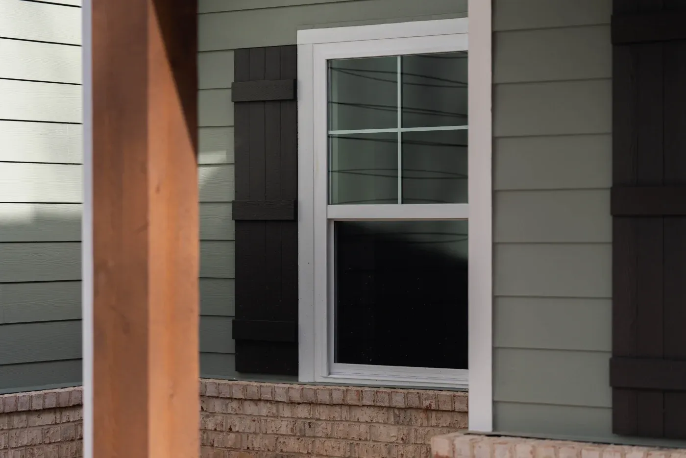 A close-up view captures a crisp white single-hung window with upper grilles set against light sage green horizontal siding, perfectly accented by dark rustic shutters and a light brick foundation base.