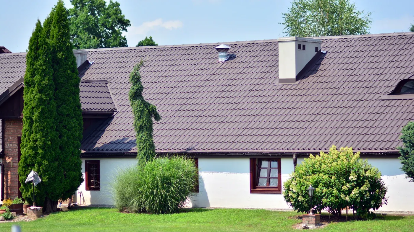 A large metal roof on a white house with windows and bushes in front.
