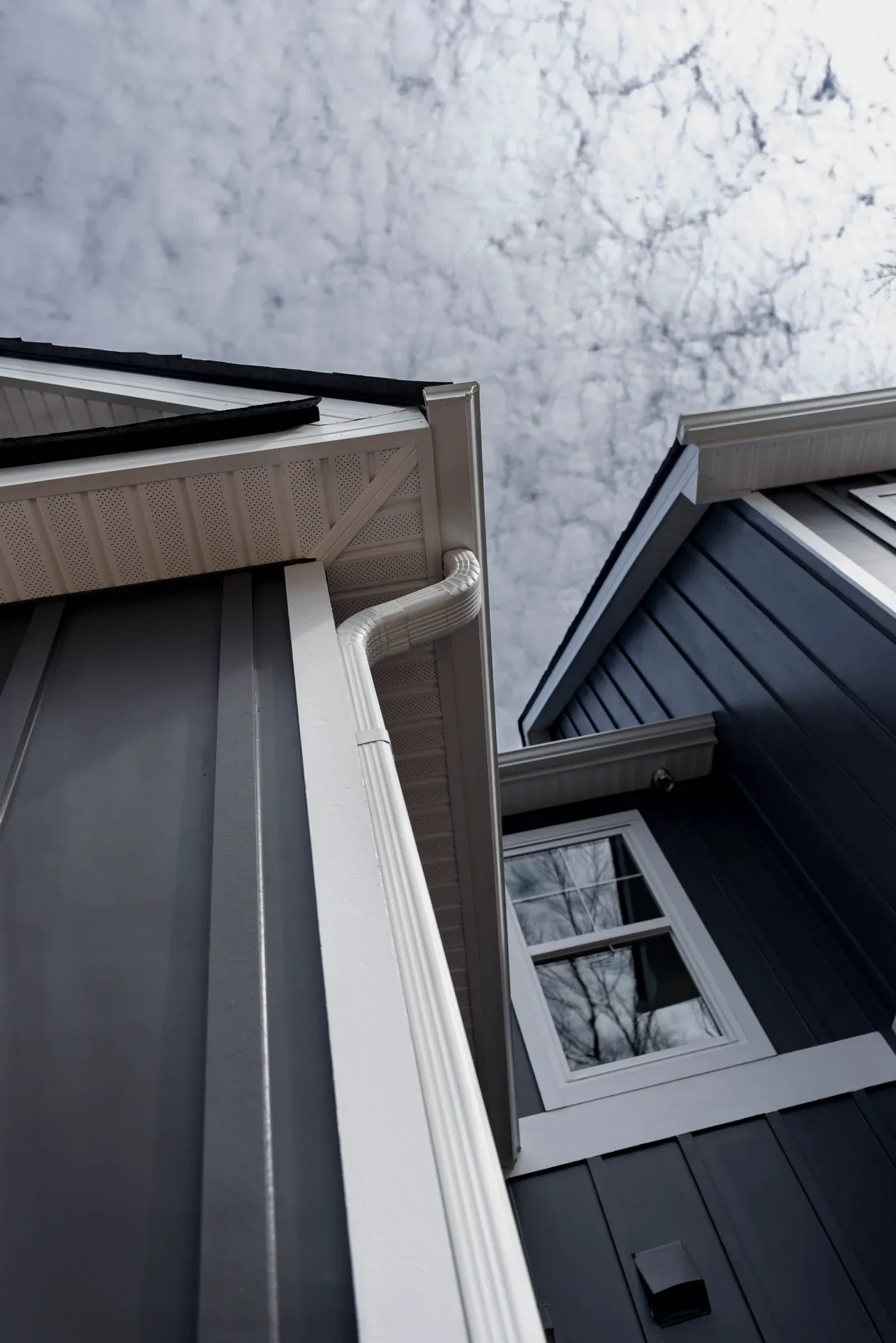 An upward angle captures a crisp white seamless gutter system and matching downspout installed neatly along a modern home featuring dark vertical board and batten siding under a cloudy sky.