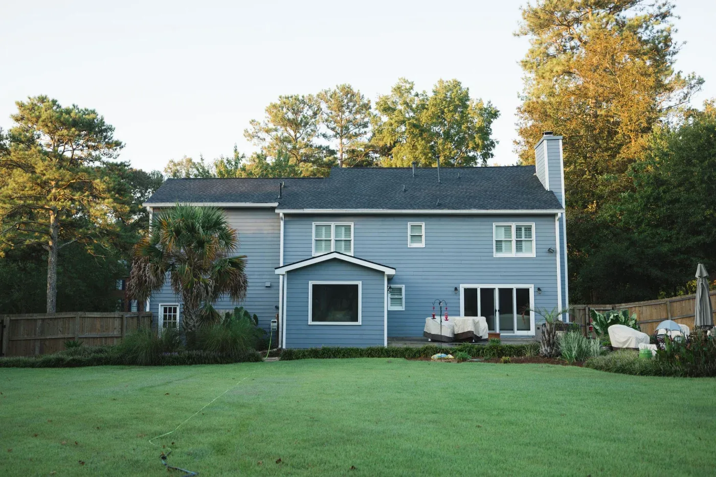 The backyard view of this two-story home highlights crisp blue-gray horizontal siding and white-trimmed windows looking out over a beautifully manicured green lawn surrounded by mature trees.
