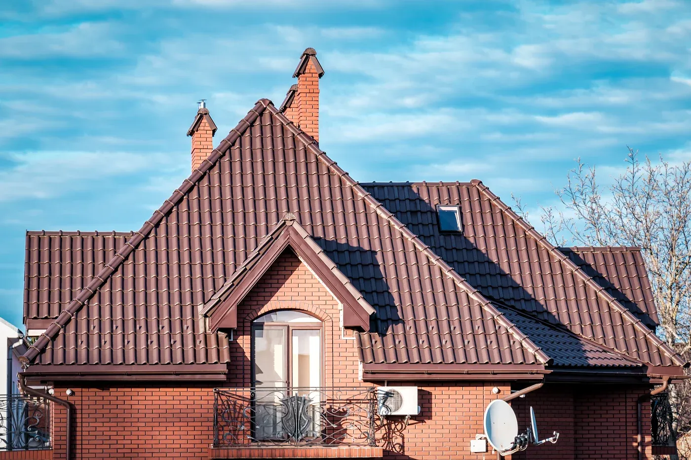 A brown metal roof on a brick house with a balcony and chimneys.