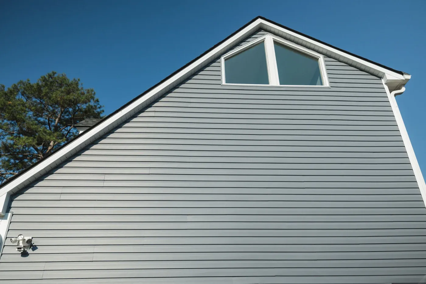 The side of a gray house with white trim and a white window near the roof.