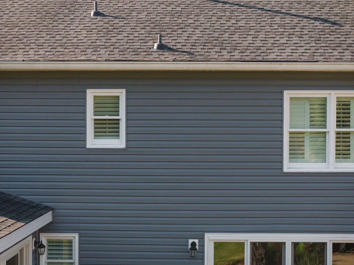 A close-up view of a gray house with white windows, white gutters, and an asphalt shingle roof.