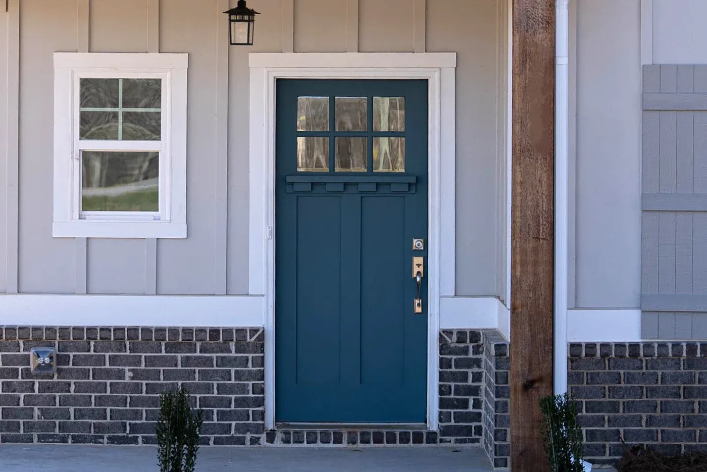A blue door with gold fixtures and glass inserts on a gray and brick house with white trim and a white window next to the door.