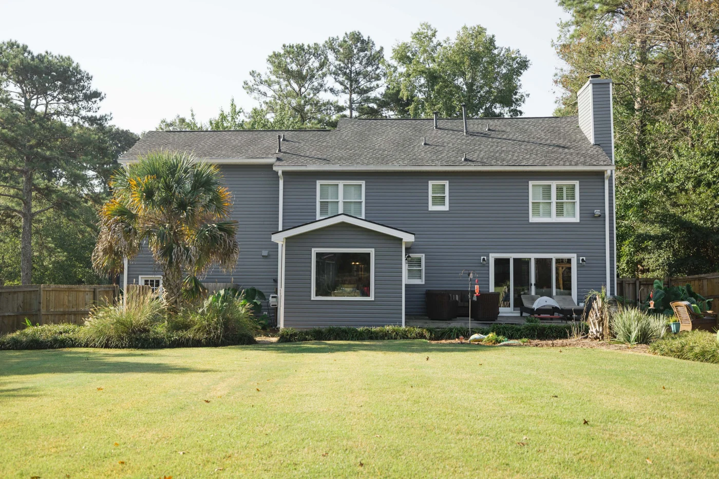A spacious backyard lawn looks up at the rear exterior of a large two-story house featuring pristine gray horizontal siding, a fenced perimeter, and professional landscaping perfect for a newly constructed home.