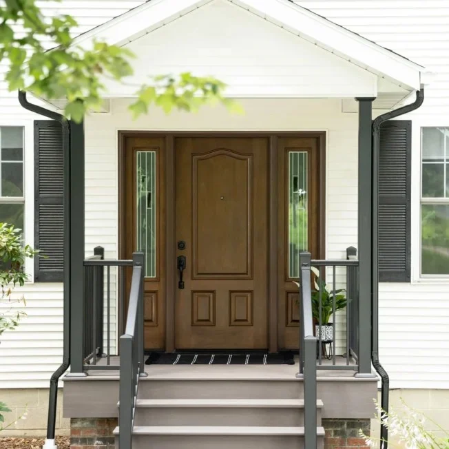 A handsome wood entry door with matching glass sidelights serves as the focal point for this welcoming front porch, beautifully framed by white horizontal siding, a gabled roof overhang, and dark railings and shutters.