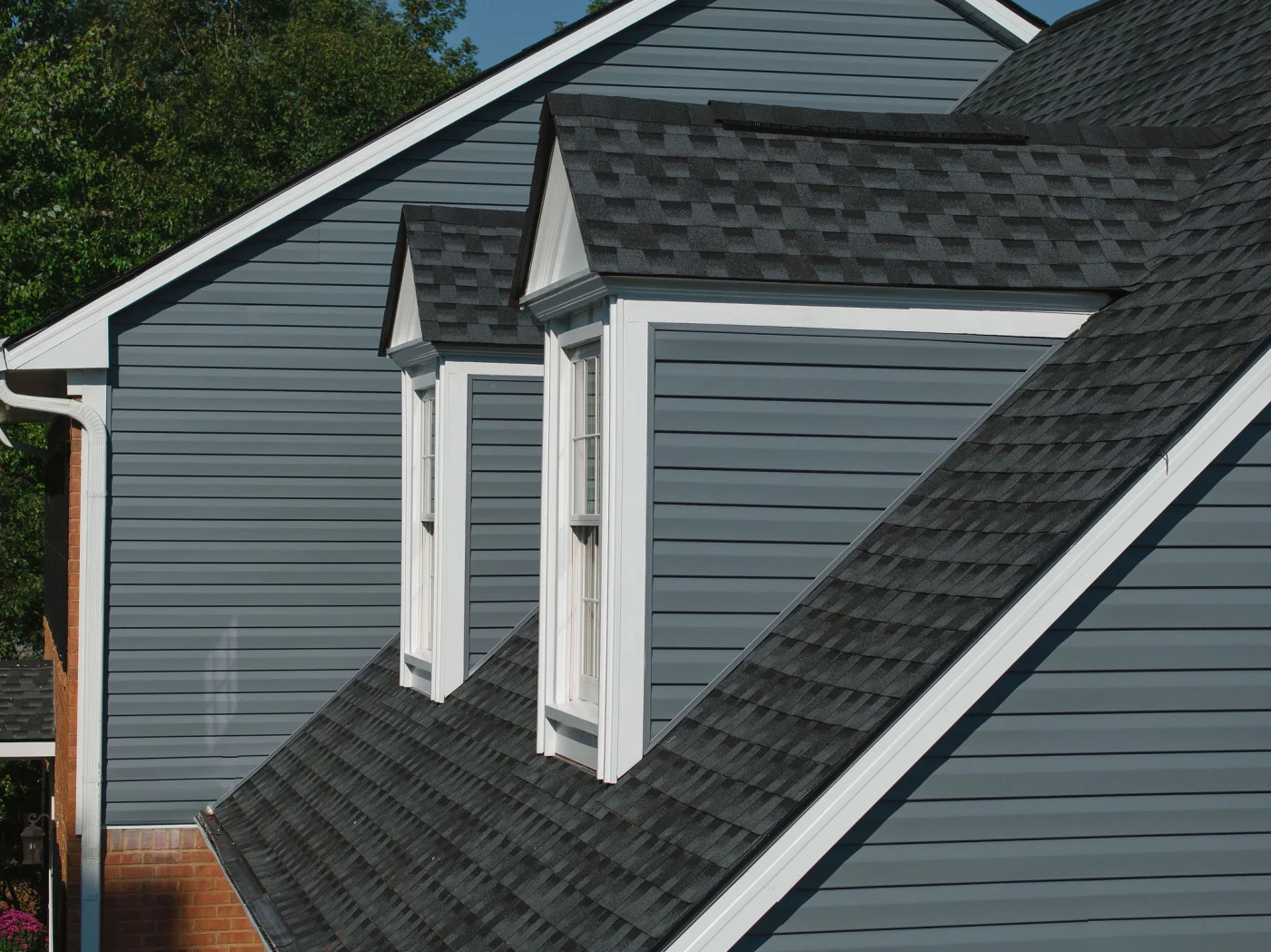 This close-up view highlights two white-trimmed dormer windows beautifully integrated into dark gray horizontal boards and a matching shingle roof, illustrating a clean and precise siding replacement.