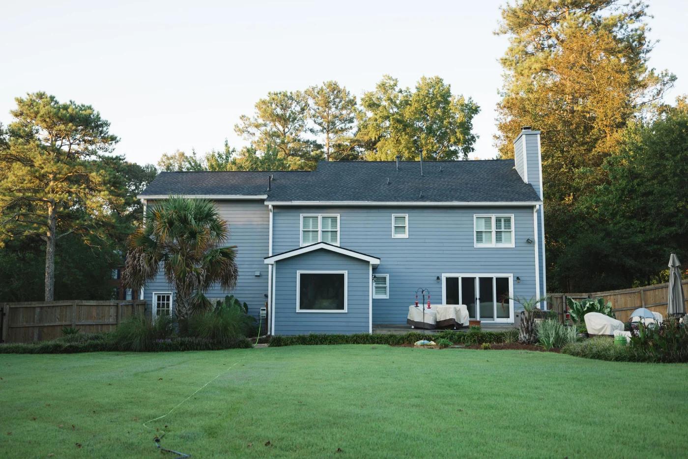 The rear exterior of this beautiful two-story home features flawlessly installed light blue horizontal siding that pairs perfectly with the manicured green lawn and gorgeous landscaping, demonstrating the exceptional quality of professional siding services.