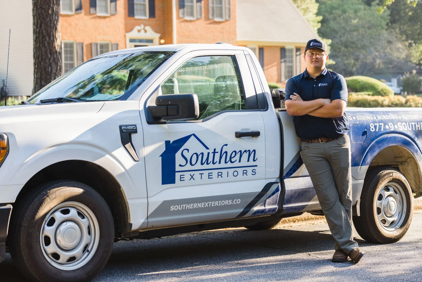 A professional wearing a branded shirt and cap stands confidently with arms crossed while leaning against a company pickup truck, representing a trusted local remodeling contractor in a residential neighborhood.