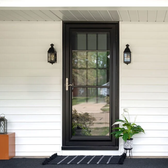 A black-framed glass storm door provides a bold, modern accent to a home front porch with white horizontal siding and symmetry from flanking lanterns, highlighting an upgrade with new windows and doors.