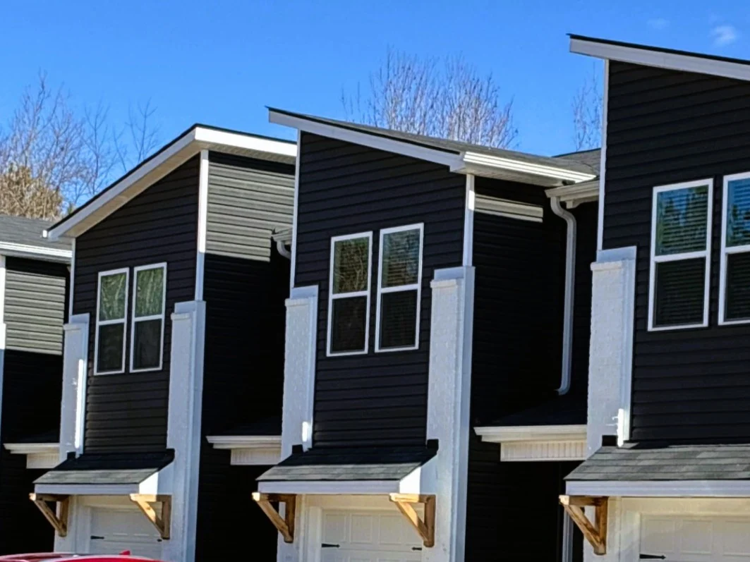 A row of modern multi-family townhomes stands out against a clear blue sky, showcasing fresh dark horizontal lap boards and crisp white vertical trim elements that highlight a complete exterior siding replacement.