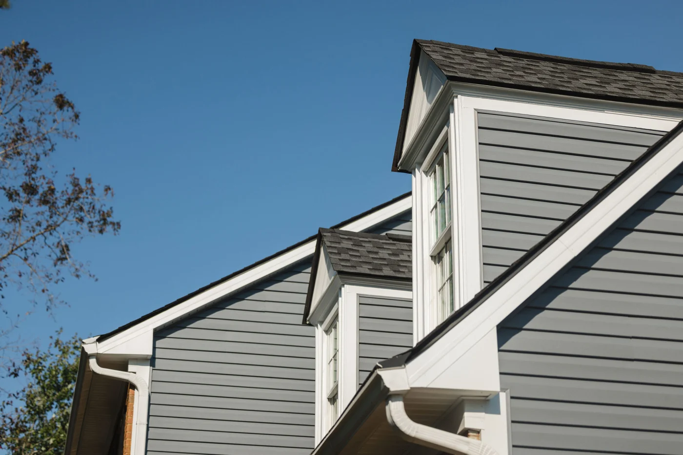 A close-up view of a home roofline highlights two gabled dormer windows with crisp white trim built into gray horizontal lap siding against a bright blue sky, showcasing expert attention to detail from professional new construction services.