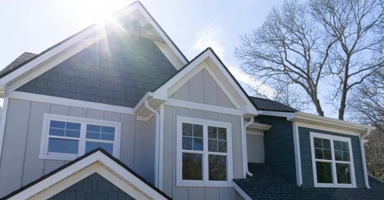 A bright sunburst illuminates a multi-gabled residential exterior featuring mixed textures of light vertical board-and-batten siding alongside dark teal shingle accents, highlighting premium quality from expert new construction services.